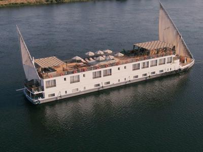 Een wit, lang rivierboot met grote zeilen en houten dek ligt op kalm water.