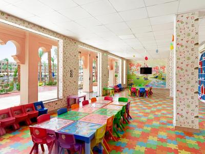 Salle de jeux colorée pour enfants avec tables, chaises et grandes fenêtres donnant sur l'extérieur.