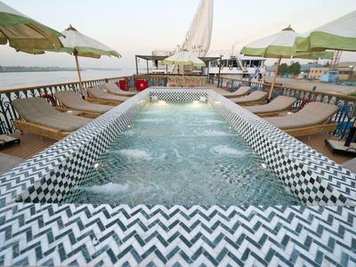 Piscine chauffée avec carreaux en mosaïque en zigzag, entourée de chaises longues et parasols au bord de l'eau.