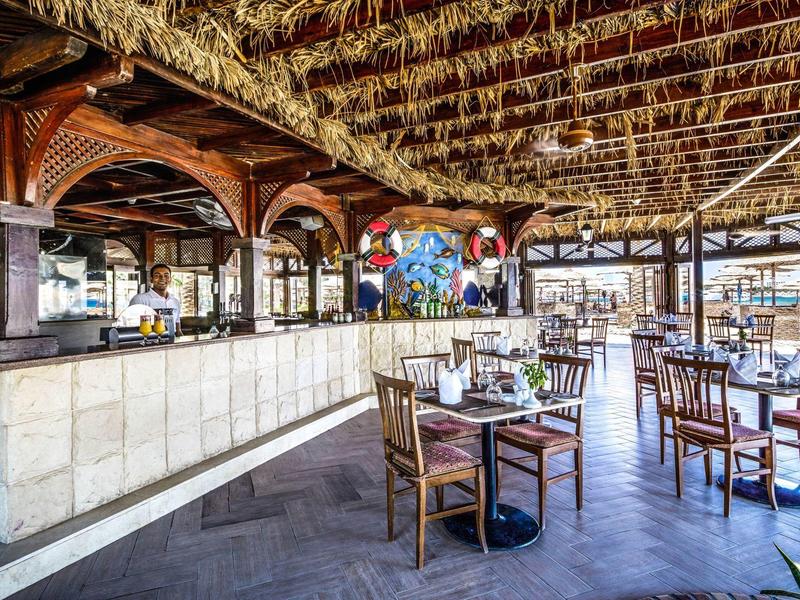 Ristorante all'aperto con tetto in paglia, tavoli e sedie in legno, atmosfera tropicale.