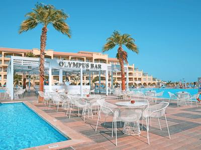 Outdoor pool with white chairs and tables under palm trees in front of Olympus Bay hotel under clear sky.