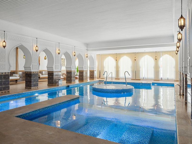 Piscine intérieure luxueuse avec colonnes et arcs traditionnels, éclairée par des lampes suspendues.