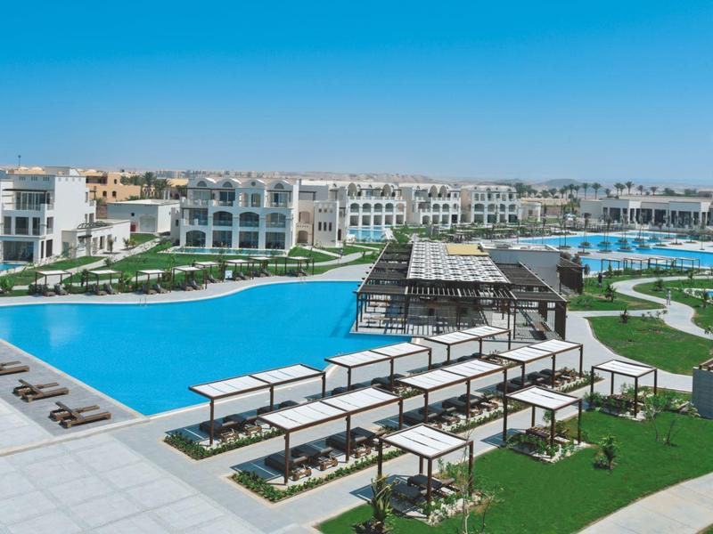 Große blaue Poolanlage mit modernen weißen Gebäuden und grünen Rasenflächen im Sonnenschein.