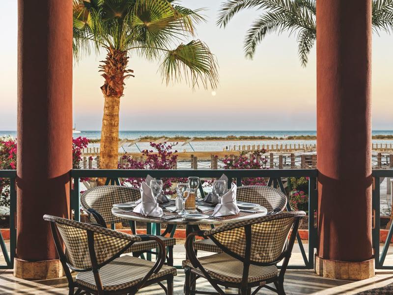 Table de restaurant extérieure avec chaises face aux palmiers et à l'océan au coucher du soleil.