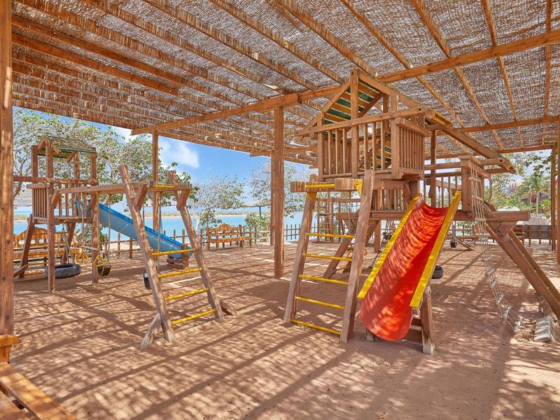 Aire de jeux extérieure avec structures en bois, balançoires, toboggan sous une pergola ombragée près de la mer.