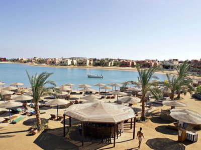 Spiaggia con ombrelloni, palme e chiosco davanti a una laguna e edifici.