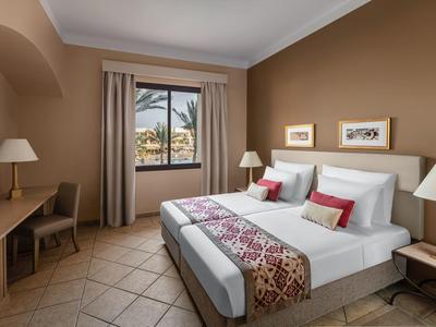 Camera d'hotel con letto matrimoniale, scrivania, lampade e vista su palme dalla finestra.