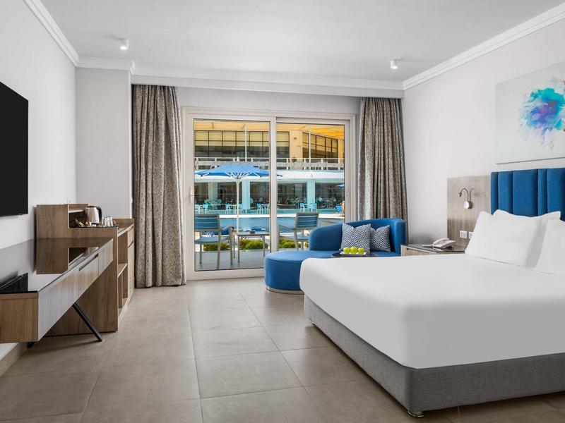 Camera d'hotel moderna con letto matrimoniale, poltrona blu e balcone con vista piscina.