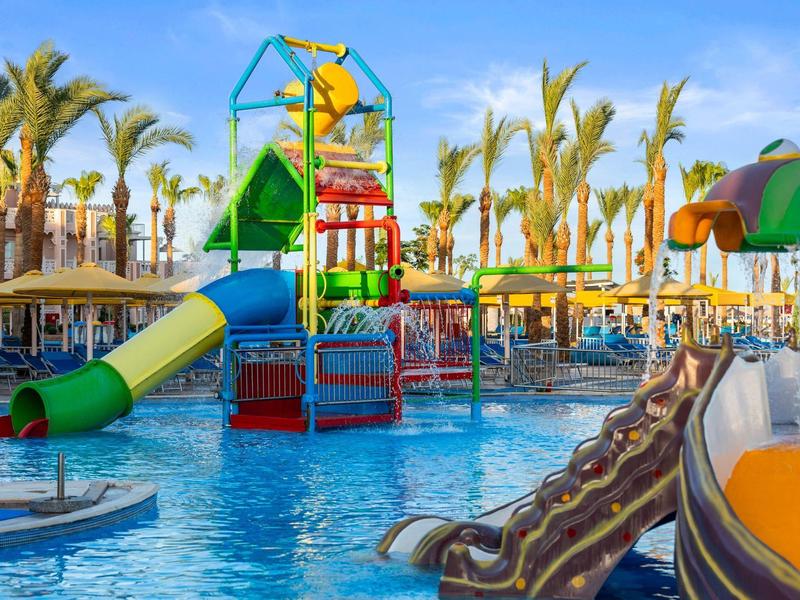 Area giochi acquatica colorata in piscina con scivoli e palme intorno.