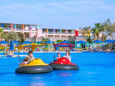 Zwei Personen fahren in bunten Bumper-Boats in einem blauen Pool vor einem Hotel.