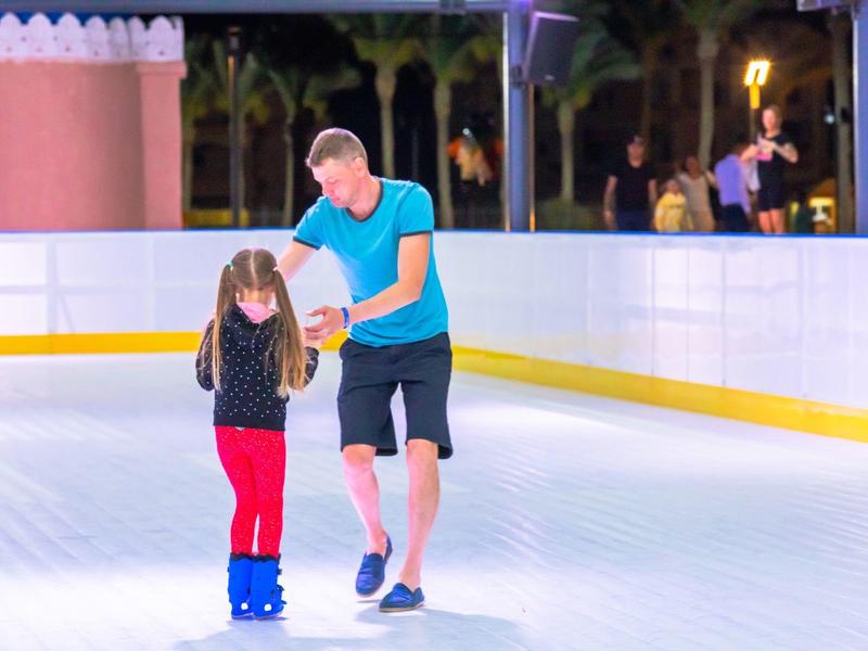 Ein Mann hilft einem Kind beim Schlittschuhlaufen auf einer Eisbahn mit Palmen im Hintergrund.