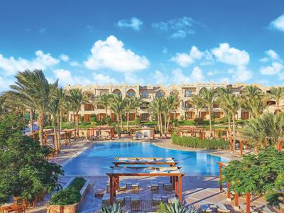 Großer Hotelpool umgeben von Palmen und mediterraner Architektur bei sonnigem Himmel.