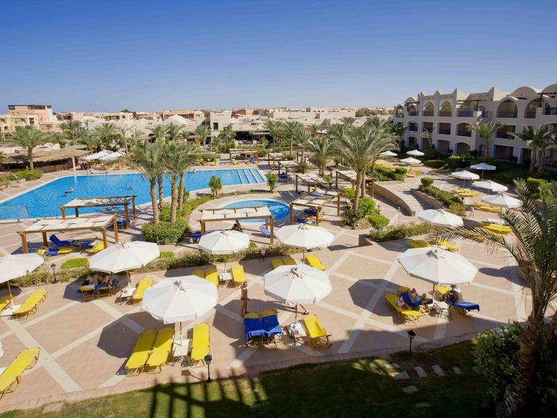 Área de piscina del hotel con sombrillas, tumbonas y edificios del hotel al fondo bajo un cielo despejado.