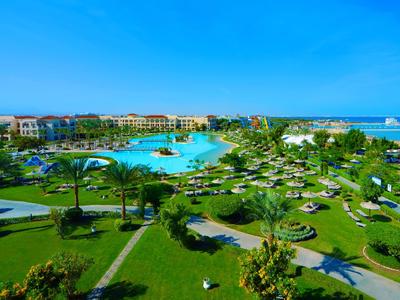 Duży kompleks hotelowy z basenem, leżakami i palmami pod czystym niebieskim niebem.