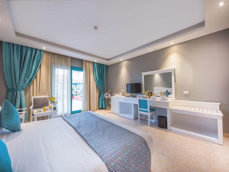 Camera d'hotel luminosa con letto, tende turchesi, scrivania e sedia vicino alla porta del balcone.