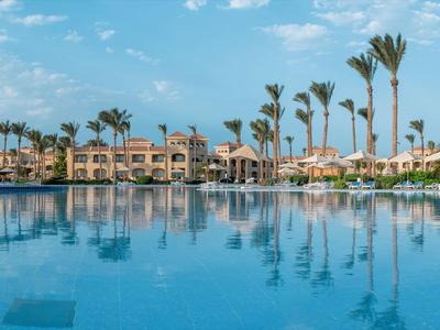 Großer Hotelpool mit Palmen vor mehrstöckigen Gebäuden unter blauem Himmel.