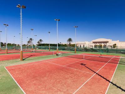 Tennis courts with red surfaces and floodlights under a clear blue sky.
