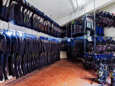A storage room with many hanging wetsuits and diving gear on white walls and red floor.