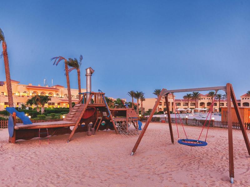 Een zand-speelplaats voor kinderen met schommel en klimrek voor een hotel onder een blauwe hemel.