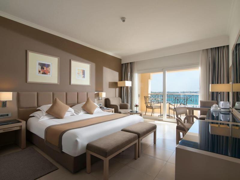 Ruime hotelkamer met groot bed, balkon, moderne meubels en zeezicht.