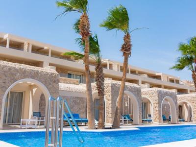 Hotel con archi in pietra e palme vicino a una piscina blu sotto un cielo soleggiato.