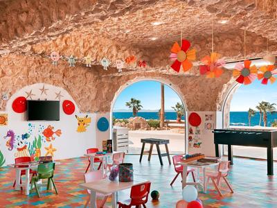 Sala giochi colorata per bambini con vista mare, tavoli piccoli, sedie e giochi sotto soffitto in pietra.