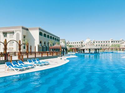 Großer blauer Pool mit blauen Liegestühlen vor einem weißen Hotelgebäude unter klarem Himmel