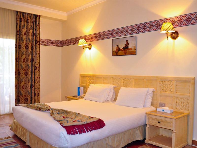 Lichte hotelkamer met tweepersoonsbed, nachtkastje, wandlampen en raam met gordijnen.