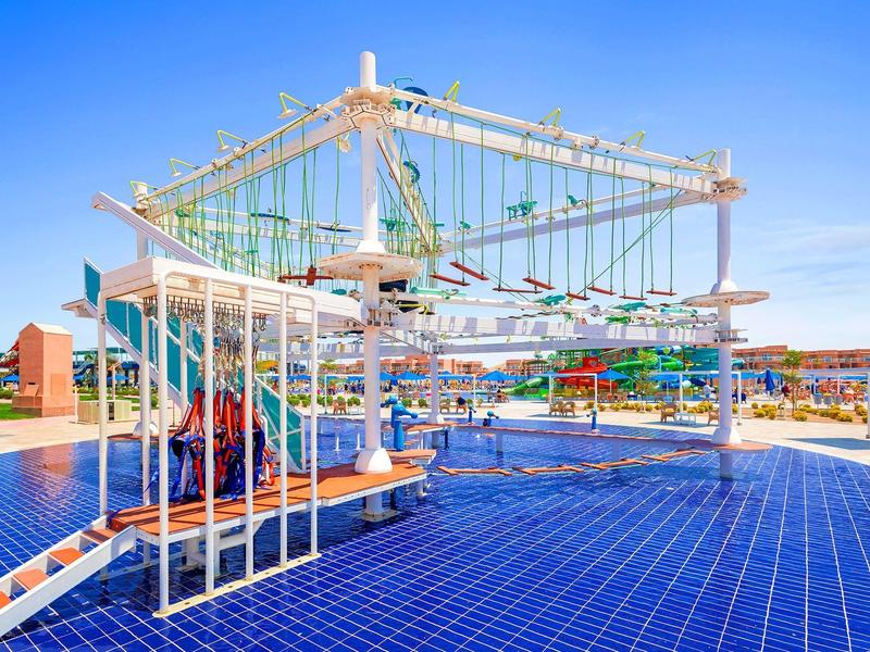 Venku dobrodružný park s lanovým parkem, horolezeckou stěnou a modrou dlažbou pod jasnou oblohou.