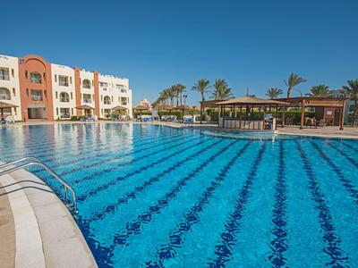 Gran piscina exterior frente a un hotel con palmeras y tumbonas bajo un cielo despejado.