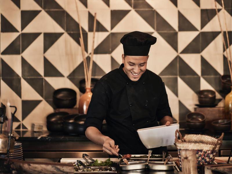 Chef preparing food in a kitchen with geometric patterned wall tiles and warm lighting.