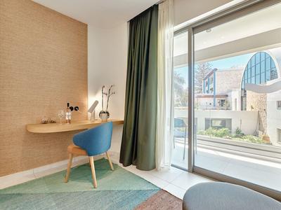 Bright hotel room with desk, blue chair, and balcony overlooking the city.