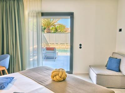 Bright hotel room with bed, armchair, blue pillow, and view of pool and loungers.