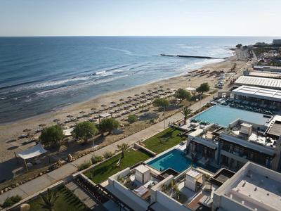 Beach with umbrellas, pool areas, and buildings along the coast under clear sky.