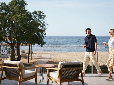 Two people walking along a beach with trees and chairs.