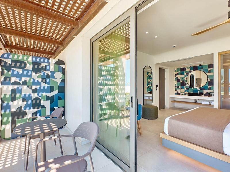Modern hotel balcony with table, chairs, and view into stylishly decorated room.