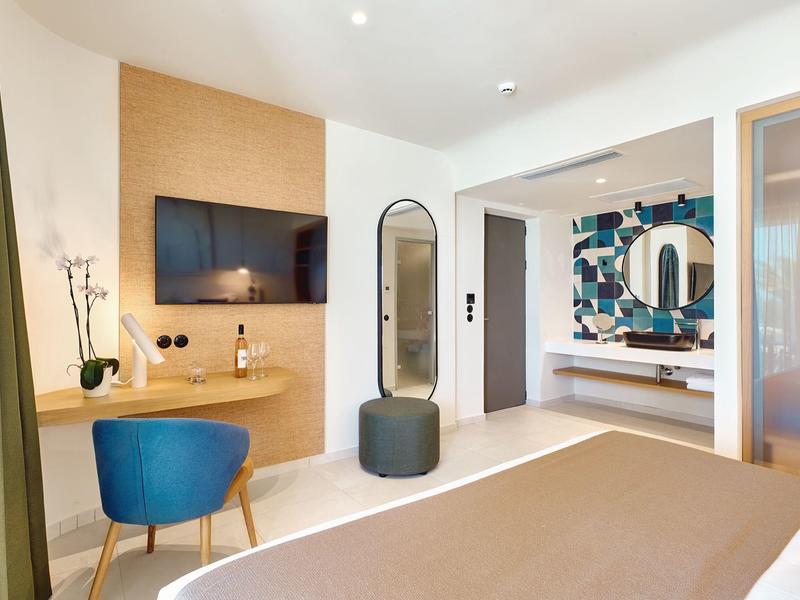 Modern hotel room with TV, blue chair, mirror, and bathroom featuring colorful tiles.