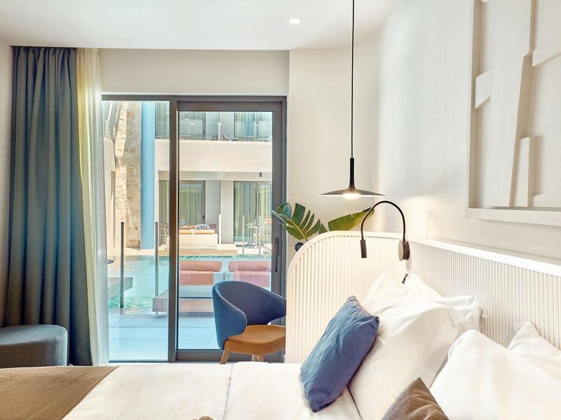 Bright hotel room with bed, armchairs, and view of terrace with pool.