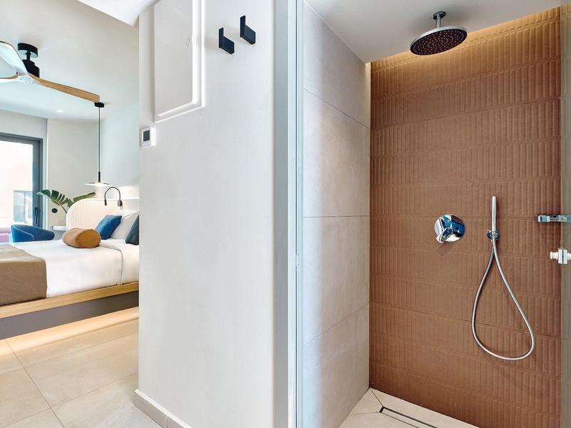 Modern hotel shower area with wooden wall adjacent to bedroom with bed and ceiling fan.