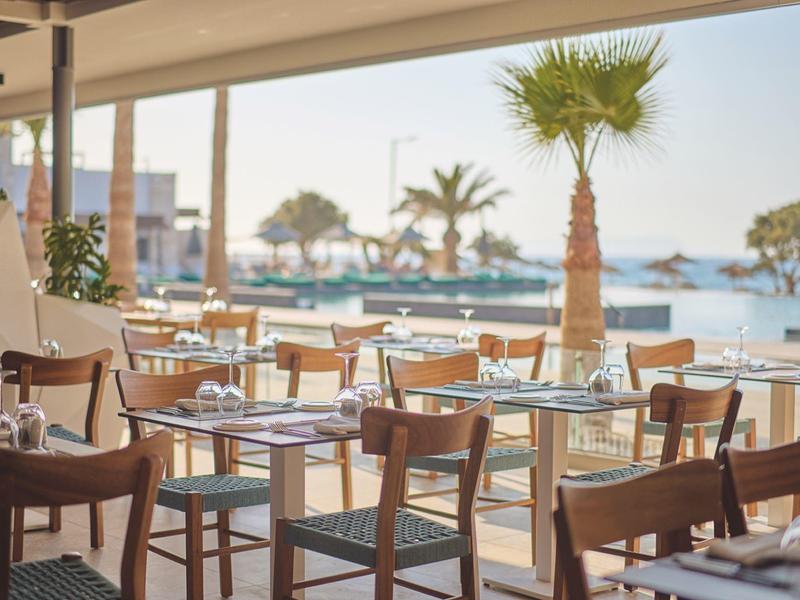 Open restaurant with wooden chairs and tables overlooking palm trees and the beach.