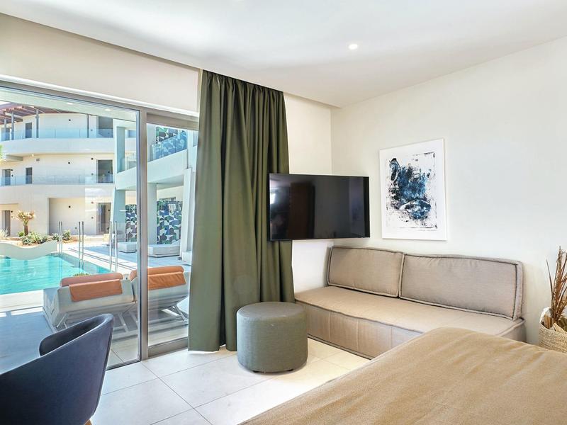 Bright hotel room with seating area, TV, and view of outdoor pool through large glass wall.