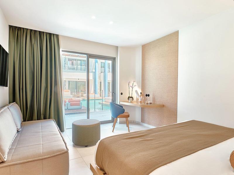 Bright hotel room with bed, sofa, desk, and view of the pool.