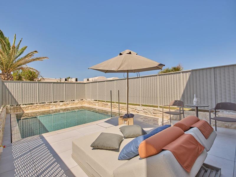 Modern pool area with lounge chair, sun umbrella, and cushions on light flooring beside a pool.