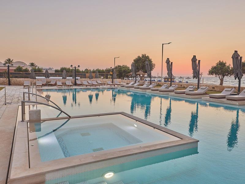 Modern hotel pool area with sun, lounge chairs, and small hot tub at sunset.