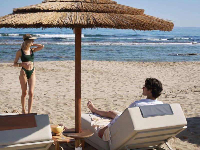 Woman in bikini stands on beach, man sits on lounge chair under thatched umbrella with sea view.