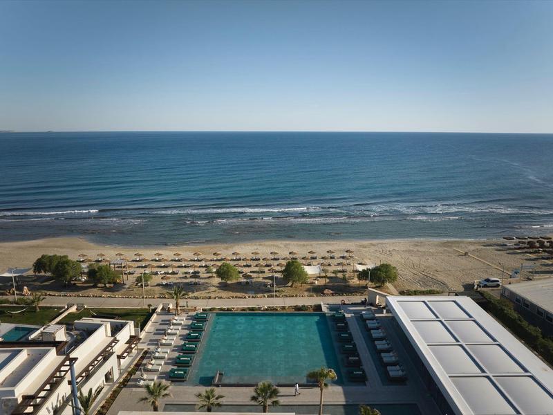 View of hotel pool, beach with lounge chairs, and calm sea under clear sky.