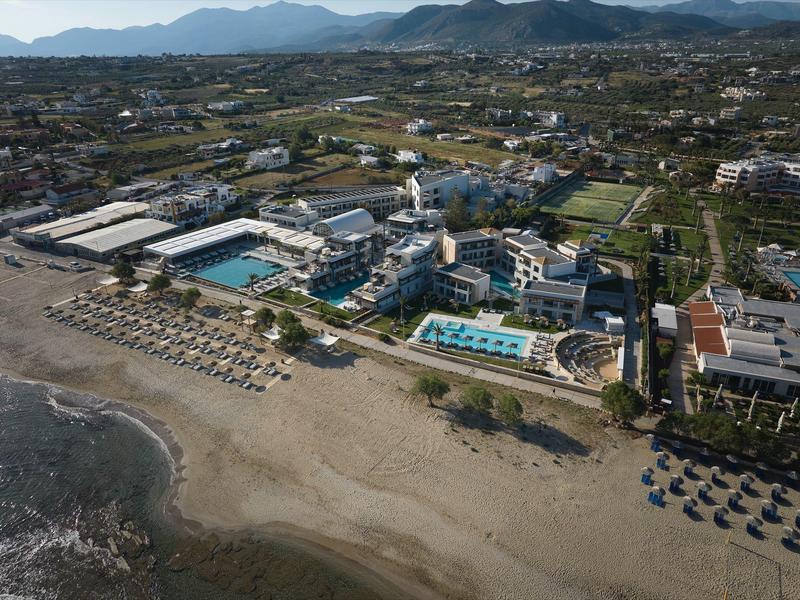 Aerial view of a hotel with multiple swimming pools on a sandy beach and mountains in the background.