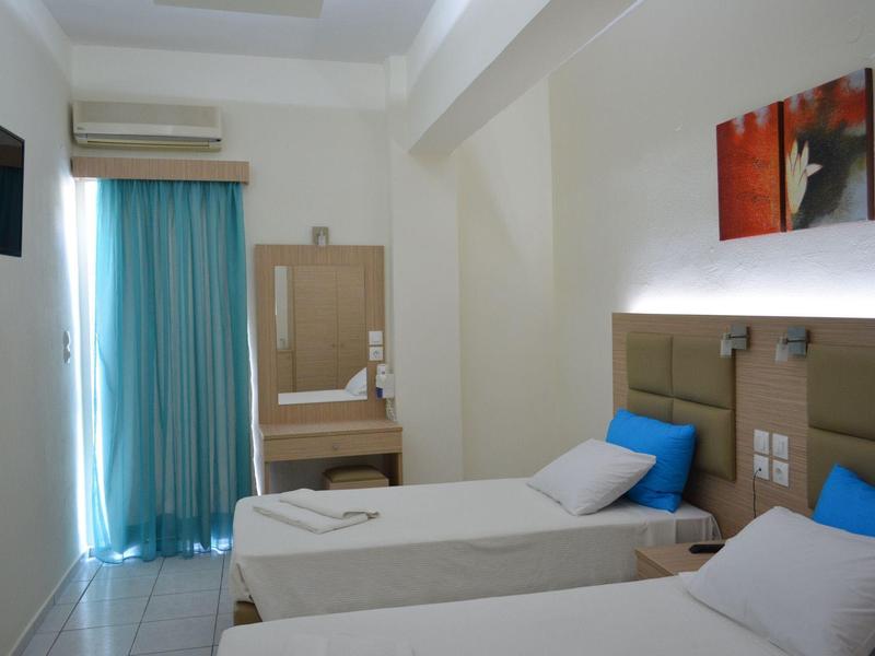 Habitación de hotel luminosa con dos camas individuales, ropa de cama azul y blanca y pequeño televisor en la pared.