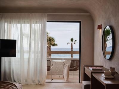 Lichte hotelkamer met gordijnen, tv en balkon met uitzicht op zee en stoelen.