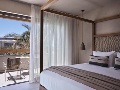 Chambre d'hôtel lumineuse avec grand lit, rideaux et balcon avec chaises et vue sur les palmiers.